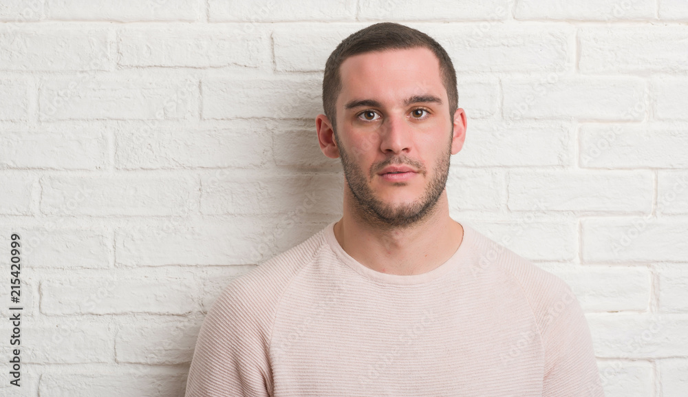 Young caucasian man standing over white brick wall with a confident expression on smart face thinking serious Young caucasian man standing over white brick wall with a confident expression on smart face thinking serious