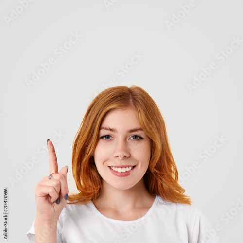 Vertical portraitof happy ginger female has pleasant smile points upwards with index finger, has freckled skin, wears casual t shirt, stands against white background with copy space. Happiness concept