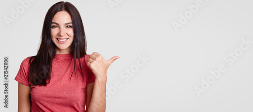 Optimistic young female with toothy friendly smile points aside with thumb, shows blank space for your advertising content or promotional text, has long dark hair, isolated over white background