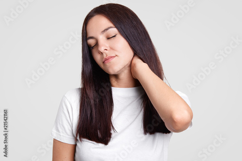 Tired beautiful brunette female keeps hand on neck, feels pain, tired after working in office, dressed in casual white t shirt, isolated over studio wall. Fatigue woman feels sick. Overworking concept