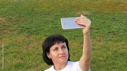 Young woman blogger sitting on the green grass in the Park and taking a selfie on her smartphone. Close-up of the girl. Technology and mobile communications. photo