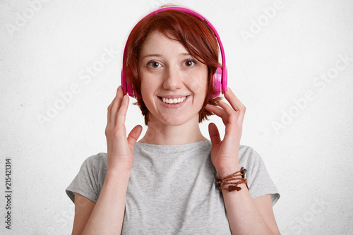 Smiling cheerful gnger femal with freckled face, listens radio or music with modern headphones, smiles broadly, dressed in casual grey t shirt, isolated over white concrete wall. Hobby concept photo