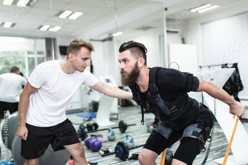 Obraz na plátně Young bearded man doing exercises in electrical muscular stimulation suit with her personal trainer at rehabilitation center