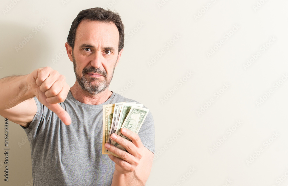 Senior man holding dollar money with angry face, negative sign showing dislike with thumbs down, rejection concept Senior man holding dollar money with angry face, negative sign showing dislike with thumbs down, rejection concept
