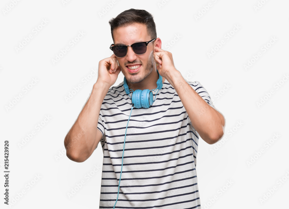 Handsome young man wearing headphones covering ears with fingers with annoyed expression for the noise of loud music. Deaf concept. Handsome young man wearing headphones covering ears with fingers with annoyed expression for the noise of loud music. Deaf concept.