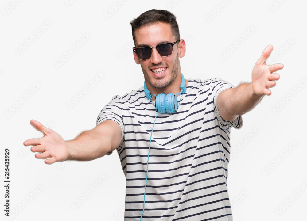 Handsome young man wearing headphones looking at the camera smiling with open arms for hug. Cheerful expression embracing happiness. Handsome young man wearing headphones looking at the camera smiling with open arms for hug. Cheerful expression embracing happiness.