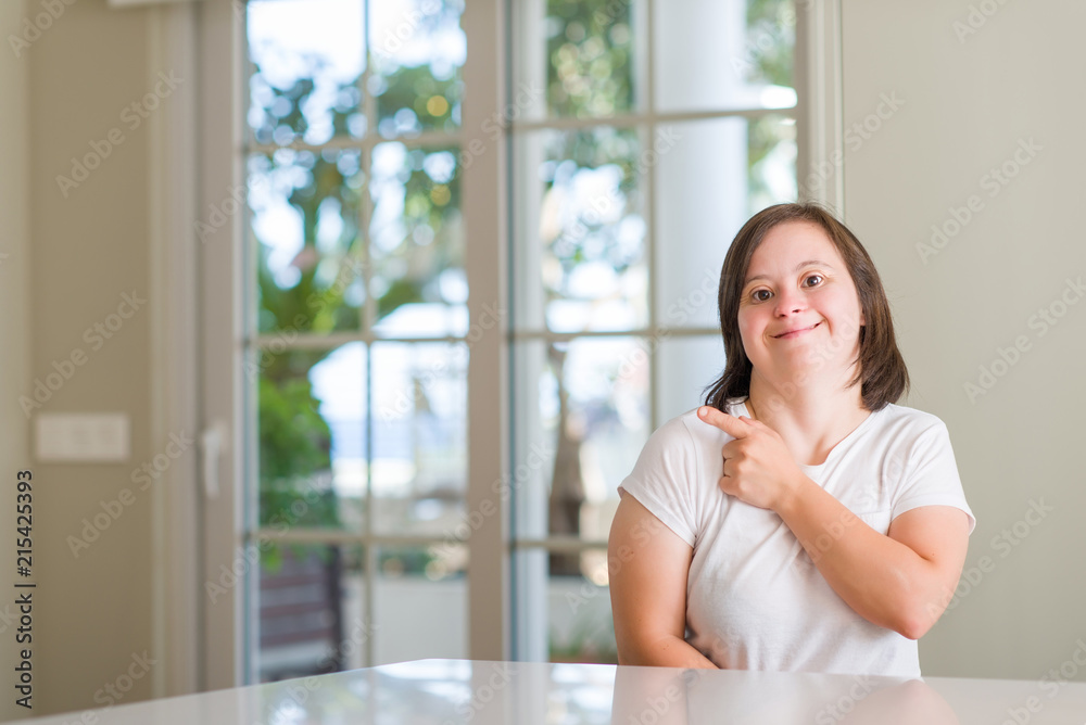 Down syndrome woman at home cheerful with a smile of face pointing with hand and finger up to the side with happy and natural expression on face looking at the camera.