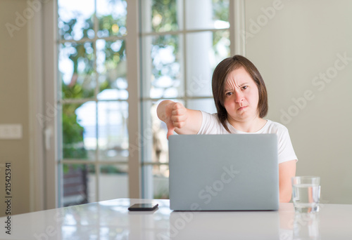 Down syndrome woman at home using computer laptop with angry face, negative sign showing dislike with thumbs down, rejection concept
