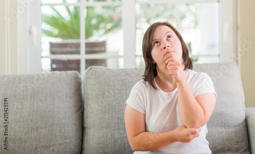 Down syndrome woman sitting on the sofa at home serious face thinking about question, very confused idea