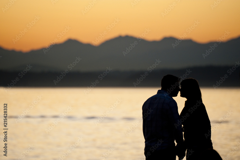 Summer Love on Alki Beach, West Seattle, Washington. A couple pauses to watch a dramatic sunset and possibly steal a kiss. The Olympic mountains are seen in the background.