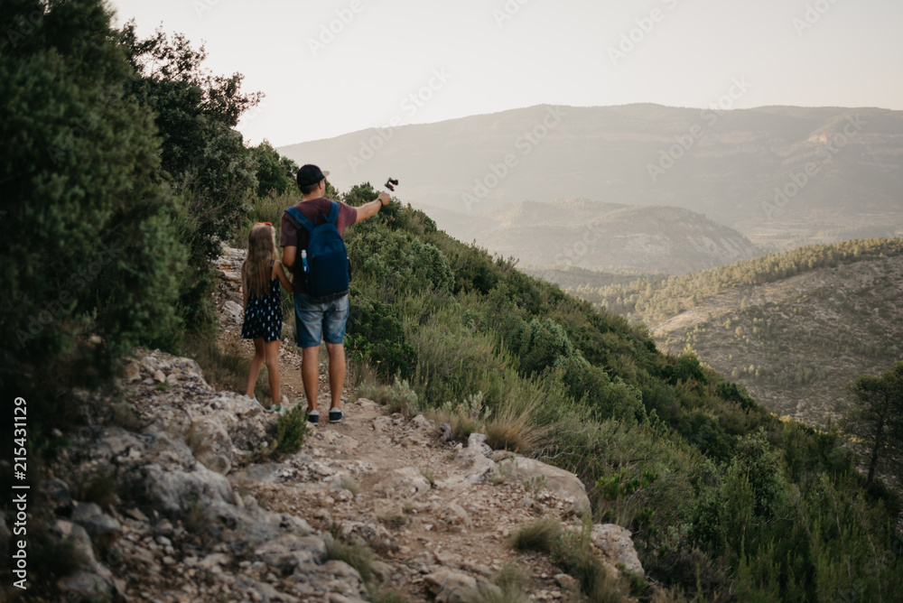 Father with daughter capturing mountains on the small camera on the sandy road on the sunset in Spain. Travelers in the mountains.