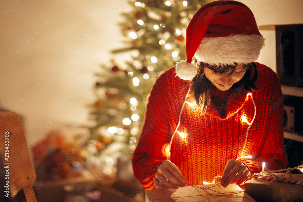 merry christmas. girl wrapping christmas presents in lights in evening festive room under tree illumination. girl in santa hat opening modern craft gifts with candy cane. atmospheric moment merry christmas. girl wrapping christmas presents in lights in evening festive room under tree illumination. girl in santa hat opening modern craft gifts with candy cane. atmospheric moment