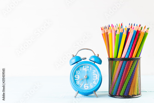 Education or back to school Concept. school office supplies. alarm clock, colorful pencils in yellow mug on white wooden background.