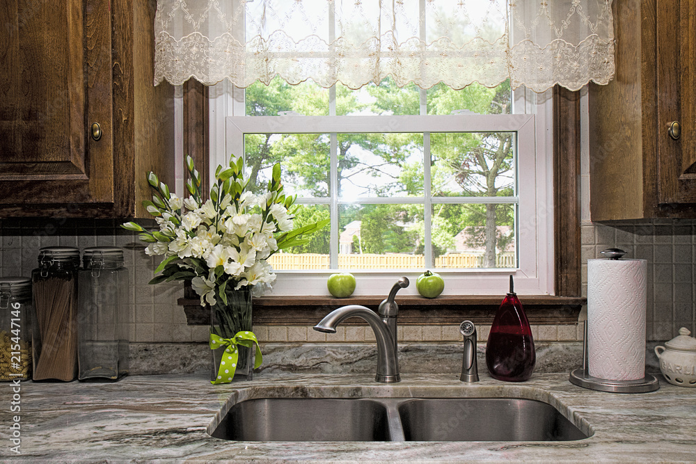 Kitchen view of window and backyard view. Sheer Valance overhead of double stainless  steel sink and granite countertop. Window seal has two green tomatoes  waiting to ripe on it. Stock Photo