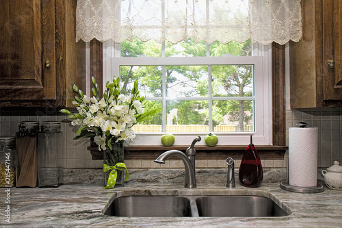 Kitchen view of window and backyard view.  Sheer Valance overhead of double stainless steel sink and granite countertop.  Window seal has two green tomatoes waiting to ripe on it.