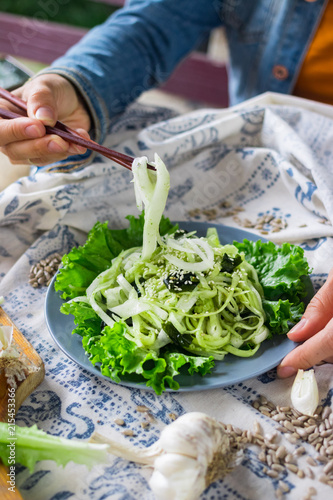 Woman hands holds and twirls zucchini spaghetti with chopsticks, raw vegan pasta with avocado sauce and salad leaves on plate. Vegan dinner, vegetarian lunch, healthy food