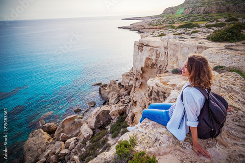 A stylish young woman traveler watches a beautiful sunset on the rocks on the beach, Cyprus, Cape Greco, a popular destination for summer travel in Europe