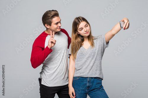 Portrait of a young couple gesture victory sign while standing together and taking a selfie isolated over gray background