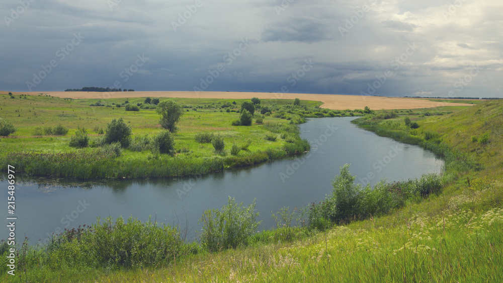 Cloudy summer landscape with fields,meadows,woods and small river.Dark stormy clouds in dramatic overcast sky.Rainy season.