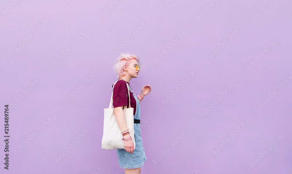Stylish girl walking around the blue wall with a shopping bag. Girl with pink hair, glasses and shopper on the background of a purple wall Stylish girl walking around the blue wall with a shopping bag. Girl with pink hair, glasses and shopper on the background of a purple wall