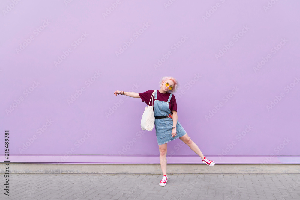 Positive stylish girl in glasses and jeans wearing a shopper in her hands against the background of a purple wall and smiling. Street portrait of a funny girl who poses on a purple background Positive stylish girl in glasses and jeans wearing a shopper in her hands against the background of a purple wall and smiling. Street portrait of a funny girl who poses on a purple background