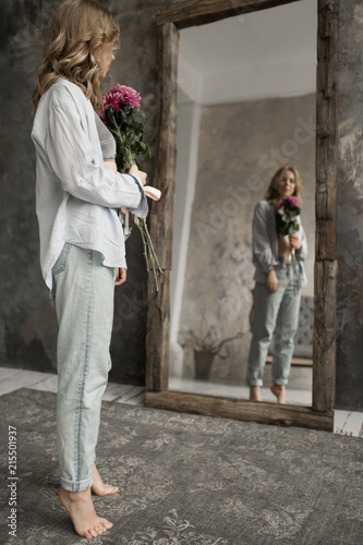 Blonde beautiful model with curly hair look in the mirror. Girl holding pink flower in studio © Anna Zenkina