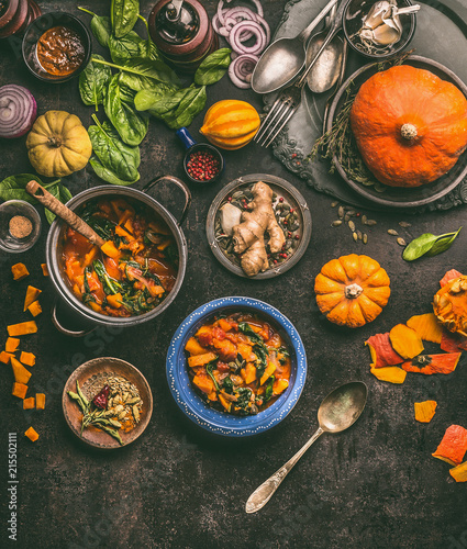 Overhead view of colorful vegetarian pumpkin stew in cooking pot and bowls with spinach and ingredients on dark rustic kitchen table background, top view. Autumn seasonal rustic country food