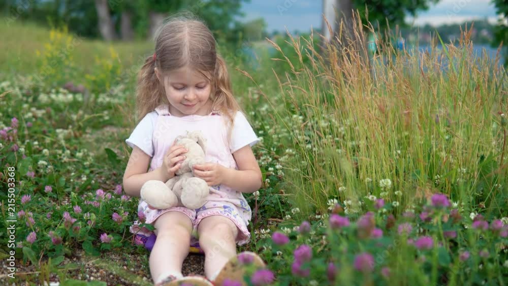 A little pretty girl plays with a plush rabbit, she sits in a meadow among a flowering clover. The child waves his hand. A little pretty girl plays with a plush rabbit, she sits in a meadow among a flowering clover. The child waves his hand.