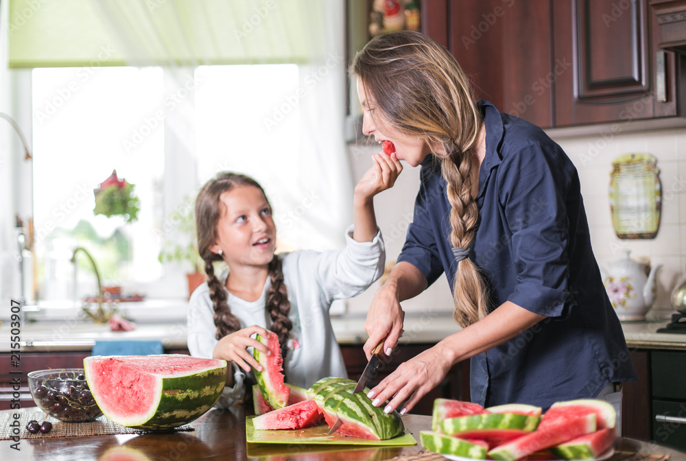 Cute little girl and her beautiful mom are cutting fruits, red watermelon and smiling while cooking in kitchen at home. Happy Family Cute little girl and her beautiful mom are cutting fruits, red watermelon and smiling while cooking in kitchen at home. Happy Family