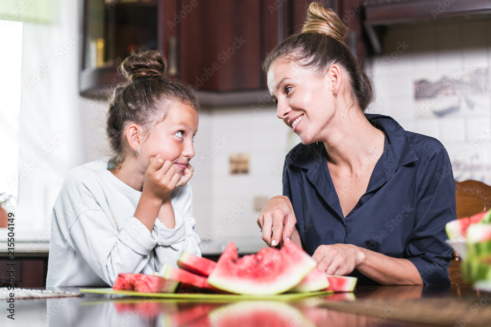 Cute little girl and her beautiful mom are cutting fruits, red watermelon and smiling while cooking in kitchen at home. Happy Family