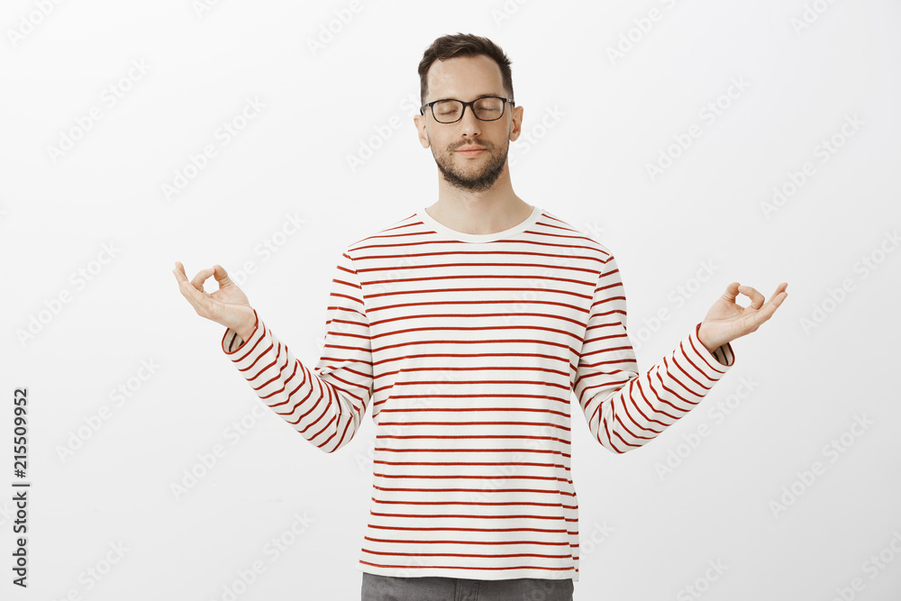 Good-looking european guy practicing yoga, wearing trendy outfit and glasses, spread hands in zen gesture and meditating with closed eyes and slight smile, feeling calm over gray background Good-looking european guy practicing yoga, wearing trendy outfit and glasses, spread hands in zen gesture and meditating with closed eyes and slight smile, feeling calm over gray background