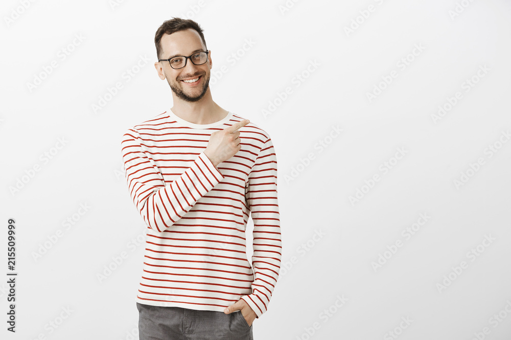 Portrat of proud confident aduly guy in glasses, holding hand in pocket and pointing at upper right corner with broad smile on face, being satisfied and joyful, feeling happy over gray background Portrat of proud confident aduly guy in glasses, holding hand in pocket and pointing at upper right corner with broad smile on face, being satisfied and joyful, feeling happy over gray background