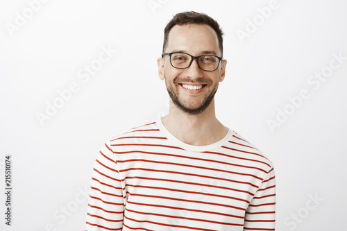 Waist-up shot of relaxed good-looking positive guy in glasses, smiling broadly and gazing at camera while talking with friend, standing against gray background, discussing interesting topic © Cookie Studio