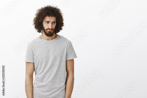 Indoor shot of handsome gloomy hispanic guy in striped t-shirt, looking from under forehead right with miserable jealous expression, feeling disappointemnt after losing something over gray background