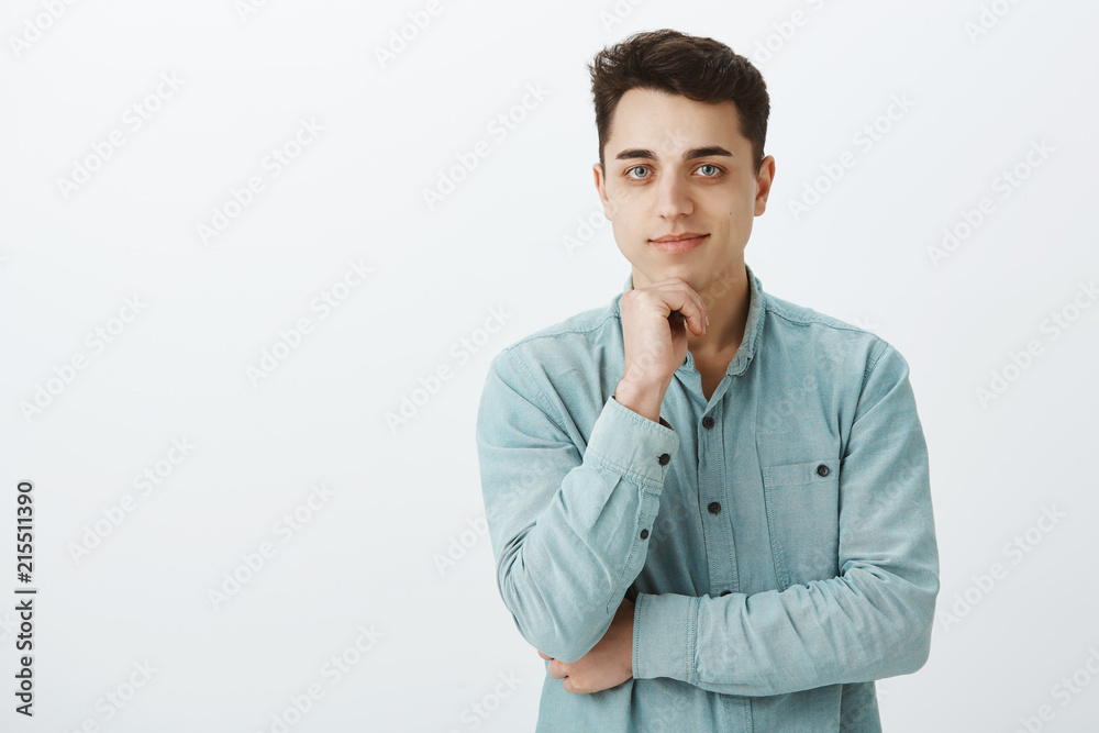 Indoor shot of curious handsome caucasian boyfriend in casual outfit, holding hand on chin and smirking at camera, thinking or making decision, being pleased and confident over gray background Indoor shot of curious handsome caucasian boyfriend in casual outfit, holding hand on chin and smirking at camera, thinking or making decision, being pleased and confident over gray background