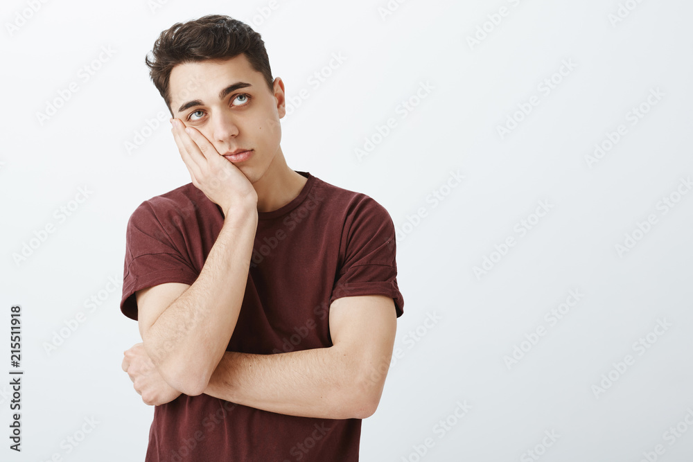 Portrait of impatient bored attractive guy in ordinary red t-shirt, leaning face on palm and looking up, waiting for end of time, being tired and feeling boredom while standing over gray background Portrait of impatient bored attractive guy in ordinary red t-shirt, leaning face on palm and looking up, waiting for end of time, being tired and feeling boredom while standing over gray background