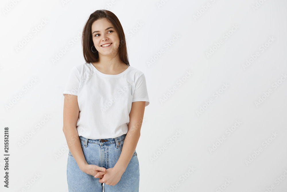 Cute stylish young woman in casual outfit, holding hands together near waist and smiling happily while gazing right, having intriguing idea in mind, dreaming and being in playful mood over gray wall Cute stylish young woman in casual outfit, holding hands together near waist and smiling happily while gazing right, having intriguing idea in mind, dreaming and being in playful mood over gray wall