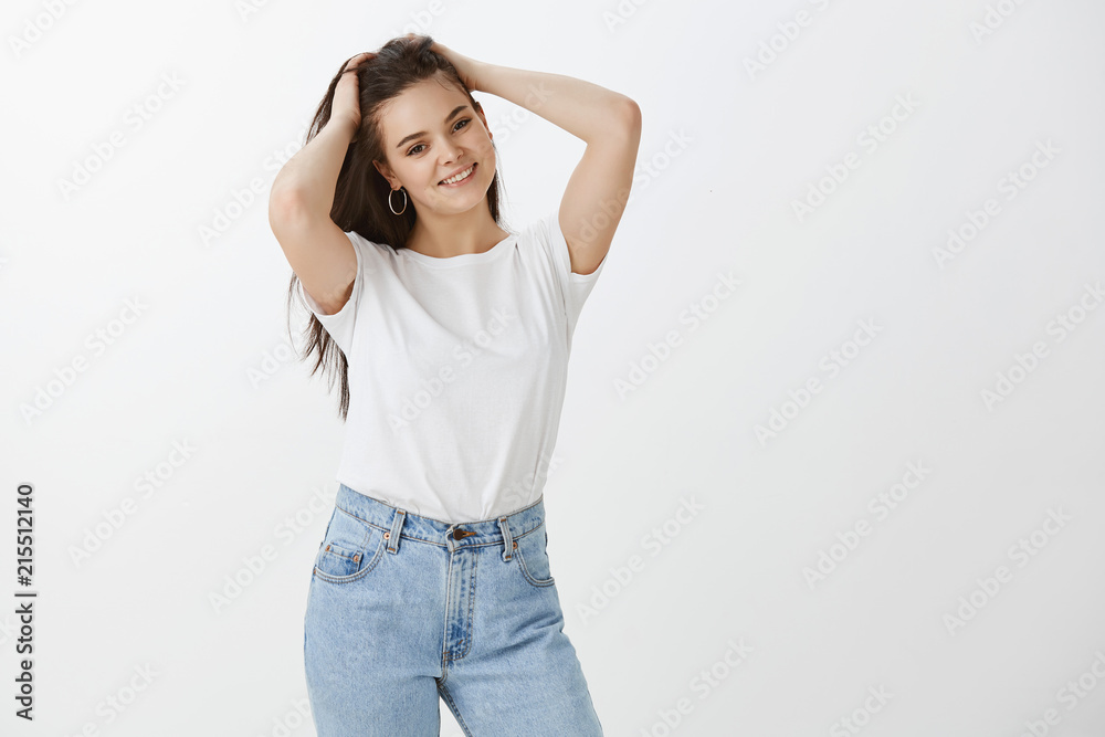 Girl asking boyfriend to take picture of her to post in internet, standing in feminine fashion pose, holding hands on hair and smiling joyfully, expressing relaxed and carefree attitude over gray wall Girl asking boyfriend to take picture of her to post in internet, standing in feminine fashion pose, holding hands on hair and smiling joyfully, expressing relaxed and carefree attitude over gray wall