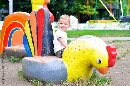 Fototapeta Naklejka Na Ścianę i Meble -  A cute two year old boy plays in a playground in a park or kindergarten. The child is sitting on the sculpture of a chicken.