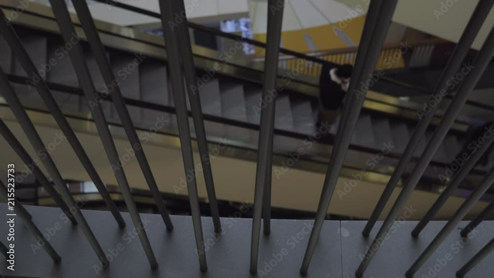 A slow motion of a moving escalator that is visible from black fence bars or some kind of railing. There is only one man standing on its steps and holding a smartphone in hands A slow motion of a moving escalator that is visible from black fence bars or some kind of railing. There is only one man standing on its steps and holding a smartphone in hands