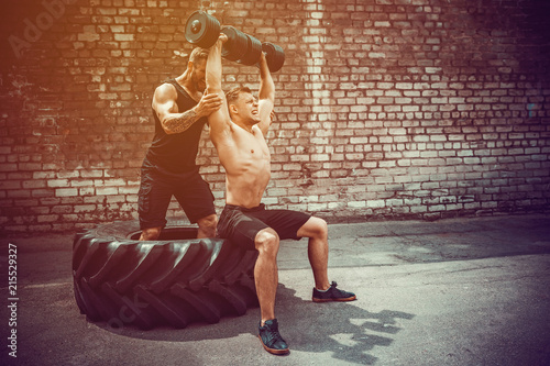 Two muscular athletes training, one raise, push the barbbell when other is motivating. Scream. Working hard. Street gym. Exercise for the shoulder muscles, deltoid.