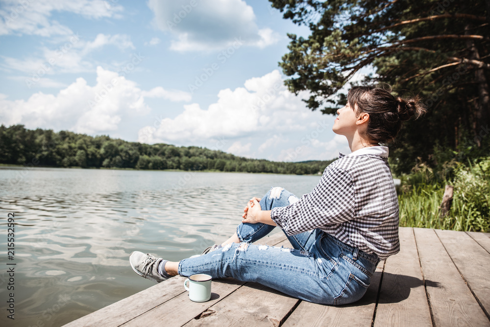 young adult woman sitting on wooden dock drinking coffee and looking at lake