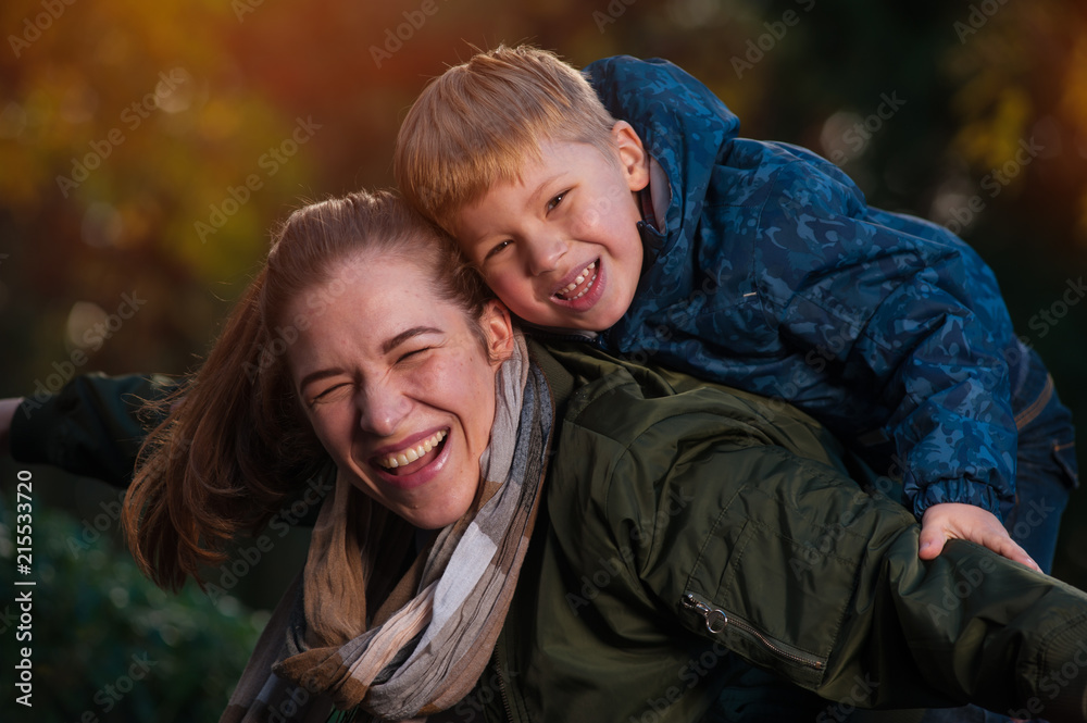 Close-up portrait - laughing positive mom and son playing together while walking in the autumn warm park Close-up portrait - laughing positive mom and son playing together while walking in the autumn warm park