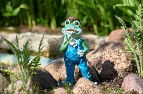 Decorative frog in the garden on the background of stones. Toy frog in overalls wipes his face with a handkerchief. photo
