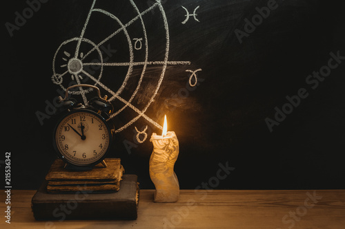 Alarm clock and old books next to a candle on a horoscope background drawn on a chalkboard