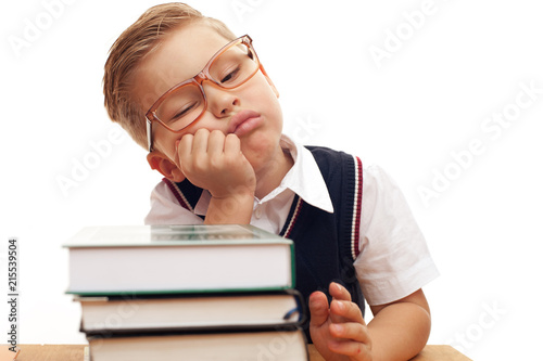 Funny sleepiing little boy wearing eyeglasses an elementary school student sitting at school desk with books on white background. Back to school. photo