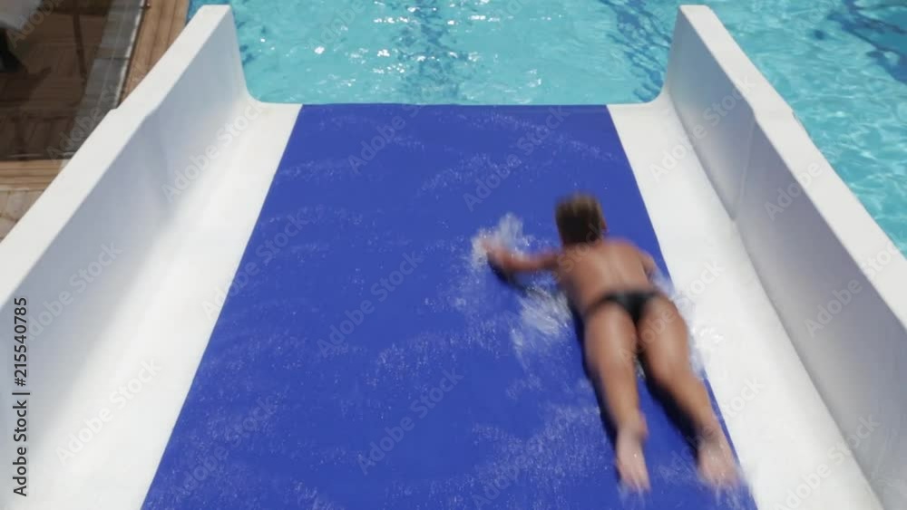 Young healthy kid cheerfully slides down blue water slide laying on belly. Real time full hd video footage.