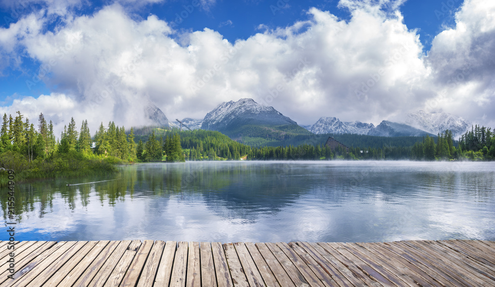 Wooden path on alpine lake Strbske Pleso with reflection of mountains, sky and clouds in the calm water, High Tatras, Slovakia Wooden path on alpine lake Strbske Pleso with reflection of mountains, sky and clouds in the calm water, High Tatras, Slovakia