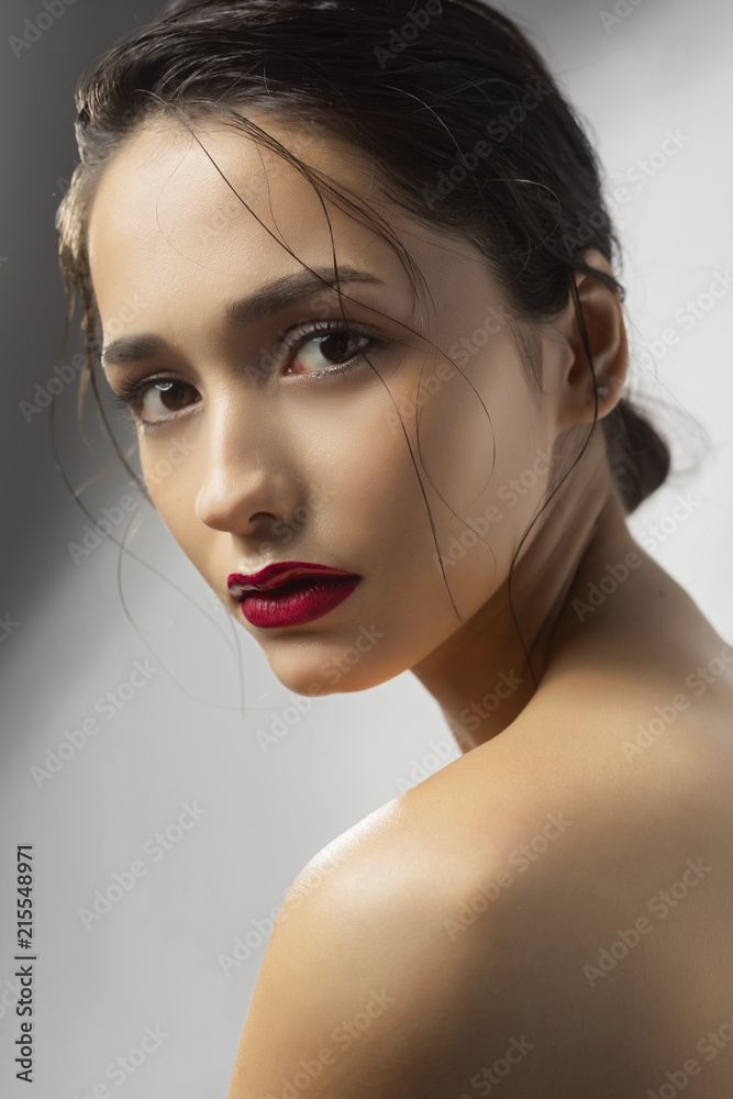 Naklejka premium Beautiful brunette girl fashion model with naked shoulders and wet hair on a gray background. Nude make-up, red lips. Healthy clean skin