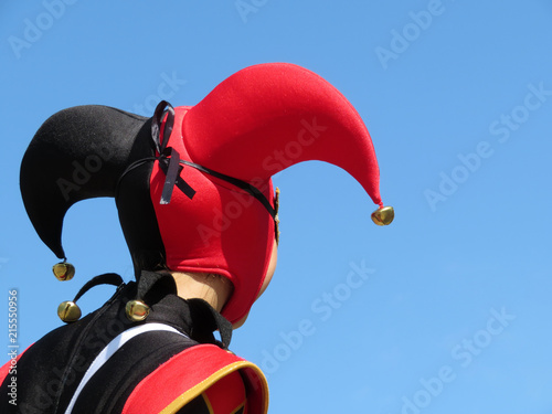Person in a jester costume isolated on clear blue sky, carnival concept. Actor in black-red foolscap with bells. Joker head close-up, back view